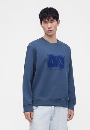 Νέος άνδρας που φοράει μπλε φούτερ Armani Exchange με λογότυπο μπροστά και ανοιχτό μπλε τζιν, σε μονόχρωμο φωτεινό φόντο.