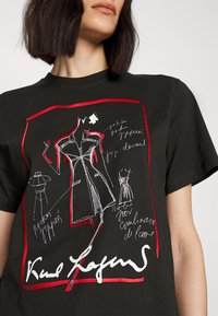 Camiseta negra con un estampado gráfico de bocetos de vestidos en blanco y rojo, con notas manuscritas. Diseño de mangas cortas y cuello redondo.