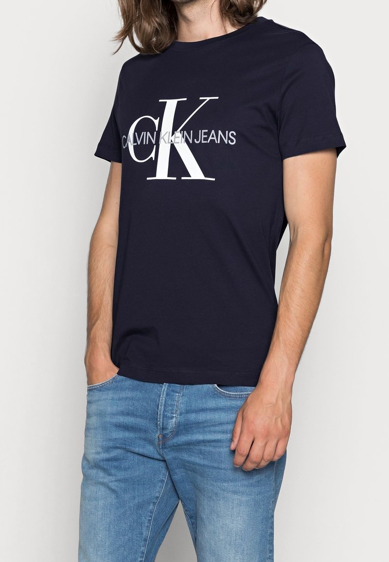 Námořnická modrá bavlněná tričko s velkým bílým logem "CK" a textem "CALVIN KLEIN JEANS", krátké rukávy a kulatý výstřih.