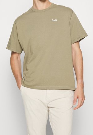 Mężczyzna ubrany w gładki oliwkowozielony t-shirt z krótkim rękawem, z małym białym napisem "forêt" na piersi, w połączeniu z jasnobeżowymi spodniami.