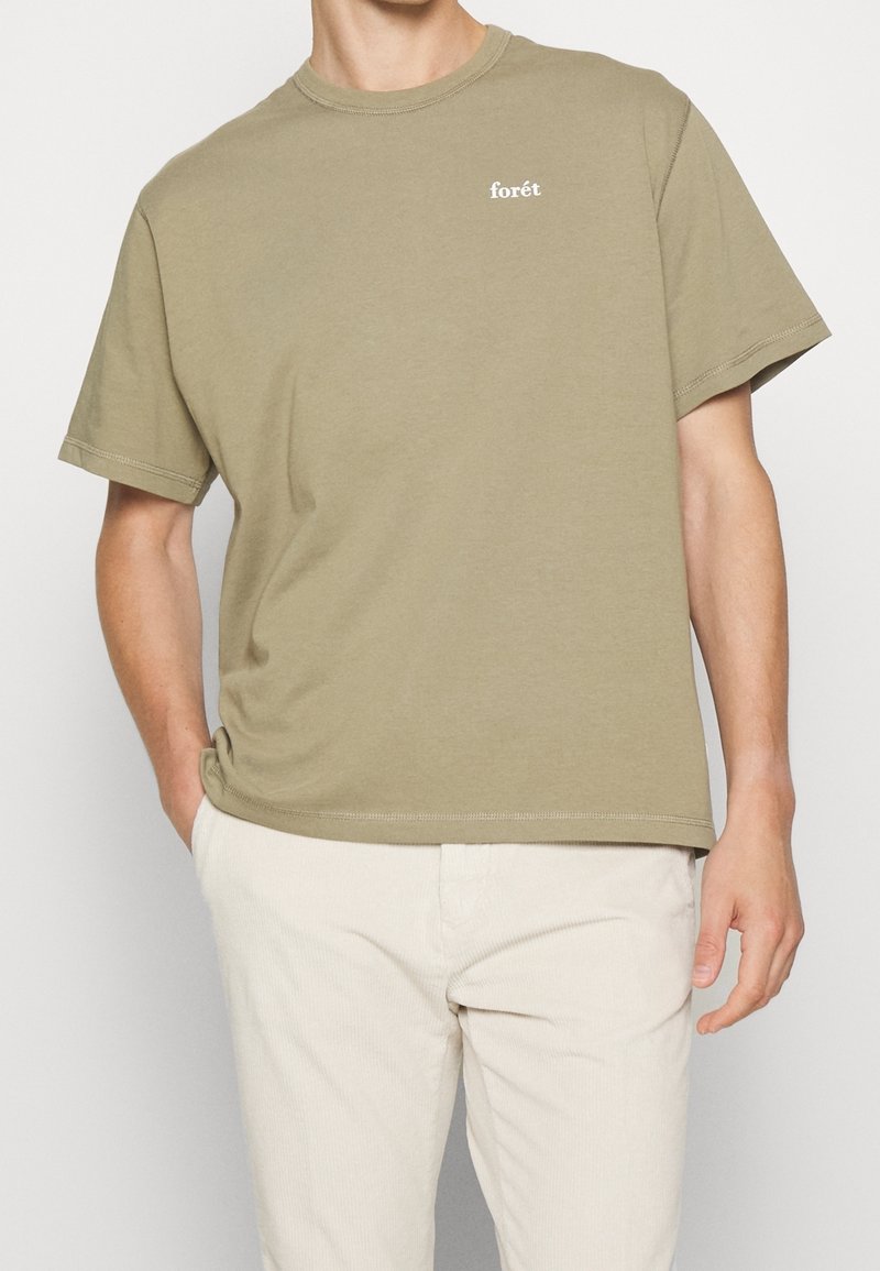 Homme portant un t-shirt à manches courtes uni vert olive avec un petit texte blanc "forêt" sur la poitrine, associé à un pantalon beige clair.