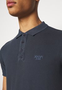 Polo shirt navy en tissu texturé, doté d'une patte de boutonnage à trois boutons et d'un logo brodé en bleu clair.