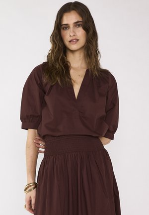 Femme aux cheveux ondulés portant un chemisier marron foncé à col en V avec des manches bouffantes et une jupe assortie taille haute froncée, debout devant un fond uni.