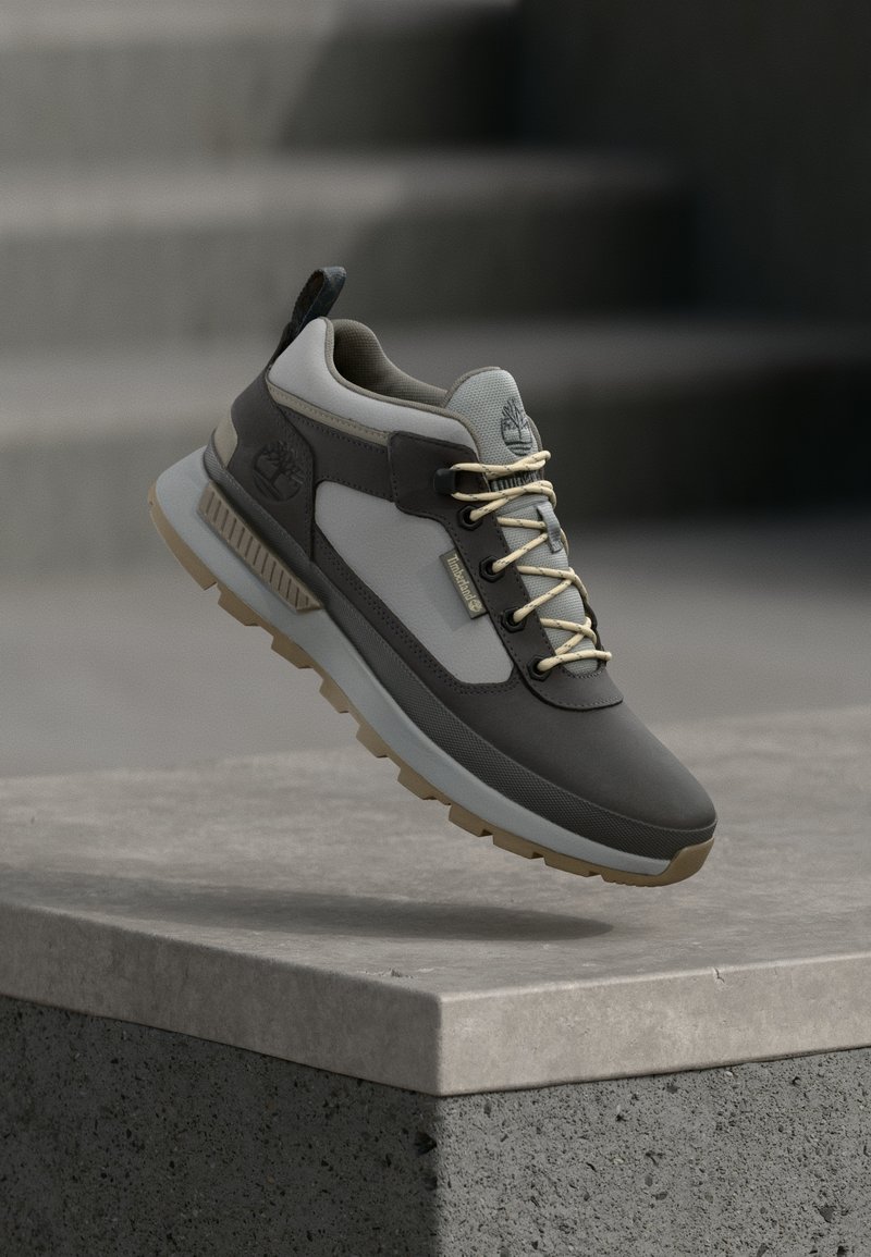 Γκρι και καφέ αθλητικό παπούτσι με υφή στην επιφάνεια, χοντρή σόλα και κορδόνια με λεπτομέρειες· το λογότυπο είναι ορατό στο πλάι και στο πίσω μέρος.