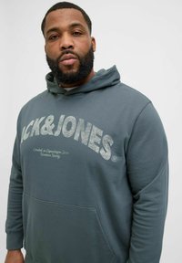 Grå hættetrøje med snoretræk i hætten, frontlomme og sølvglitrende logo tekst "JACK & JONES." Glat stoftekstur og afslappet pasform.