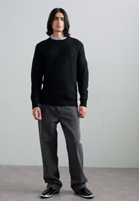 Pulover negru din tricot cu textură, cu manșete din knit și tiv zimțat, asortat cu blugi gri largi și încălțăminte neagră, pe un fundal neutru.