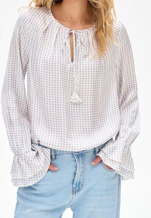 Femme portant un chemisier blanc à pois bleu clair, col noué, poignets à volants et jean en denim bleu clair, mains dans les poches.