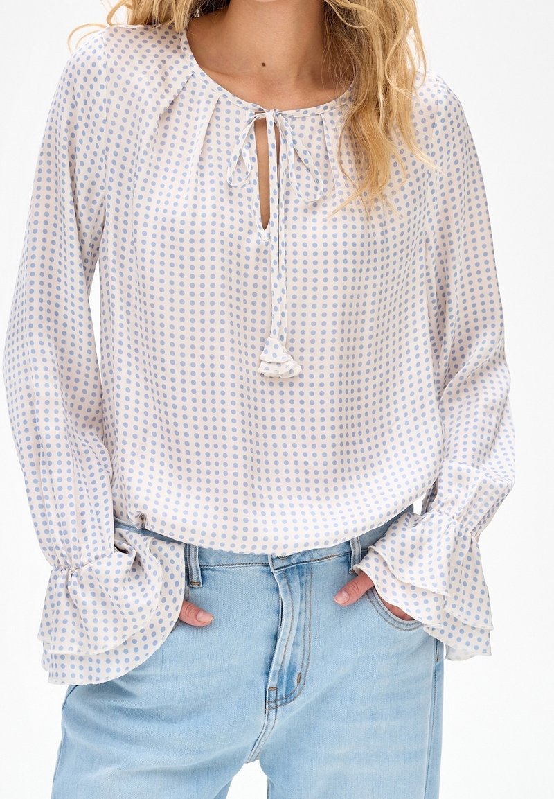 Femme portant un chemisier blanc à pois bleu clair, col noué, poignets à volants et jean en denim bleu clair, mains dans les poches.