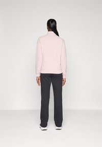 Sweatshirt rose clair avec un col montant, associé à un pantalon de survêtement gris foncé. Texture lisse, design simple et coupe décontractée.