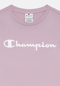 T-shirt en coton violet clair avec un col rond et un logo "Champion" blanc bien visible imprimé sur le devant. Étiquette de soin attachée.