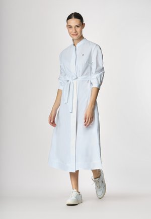 Abito bianco in cotone, con cintura, maniche lunghe, chiusura a bottoni e un sottile dettaglio del logo sul petto. Abbinato a sneakers bianche.