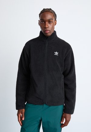 Black fleece jacket with high collar and full zipper; features white Adidas logo on the left chest; soft texture, oversized fit.