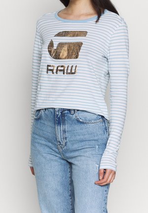 Femme portant une chemise à manches longues rayée bleu clair et blanc avec un logo doré métallique "RAW", associée à un jean taille haute bleu clair.