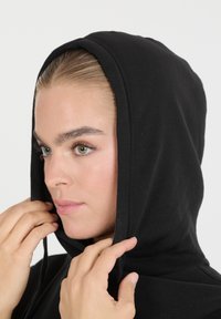 Nuori henkilö säätää mustaa huppariaan, jolla on vaalea iho, vihreät silmät ja vaaleanruskea hius, joka on vedetty taakse, yksinkertaista valkoista taustaa vasten.