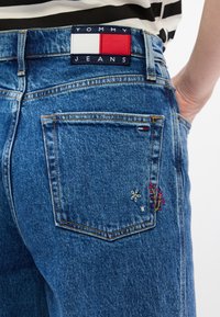 Zili džinsa bikses ar "Tommy Jeans" uzlīmi, aizmugurējā kabatā izšuvumi ar ziedu rakstiem rozā un baltā krāsā, kā arī redzama šūšana.