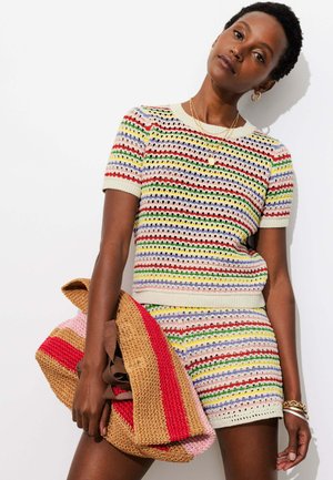 Femme portant un haut en maille multicolore à rayures à manches courtes et un short assorti, tenant un grand sac tissé rayé rouge, rose et beige.