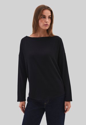 Mujer con cabello castaño claro, lleva una blusa negra de manga larga y suelta y vaqueros azul oscuro, de pie frente a un fondo gris liso.