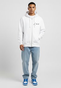 Felpa bianca con zip e coulisse, testo stampato nero, tasche anteriori. Abbinata a jeans blu chiaro e sneakers blu e bianche.