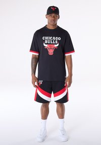 Fekete póló "CHICAGO BULLS" felirattal és logóval, piros-fehér csíkos ujjakkal. Fekete rövidnadrág piros-fehér részletekkel. Fehér tornacipő.