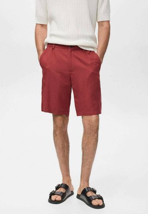 Pantaloni corti rossi in cotone con una vestibilità rilassata, dotati di passanti per la cintura e tasche laterali, abbinati a sandali neri con una fibbia decorativa.