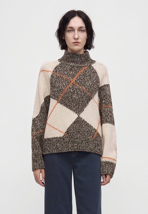Woman with short dark hair wearing a high-neck sweater with brown, beige, and orange geometric patterns and dark pants against a plain background.