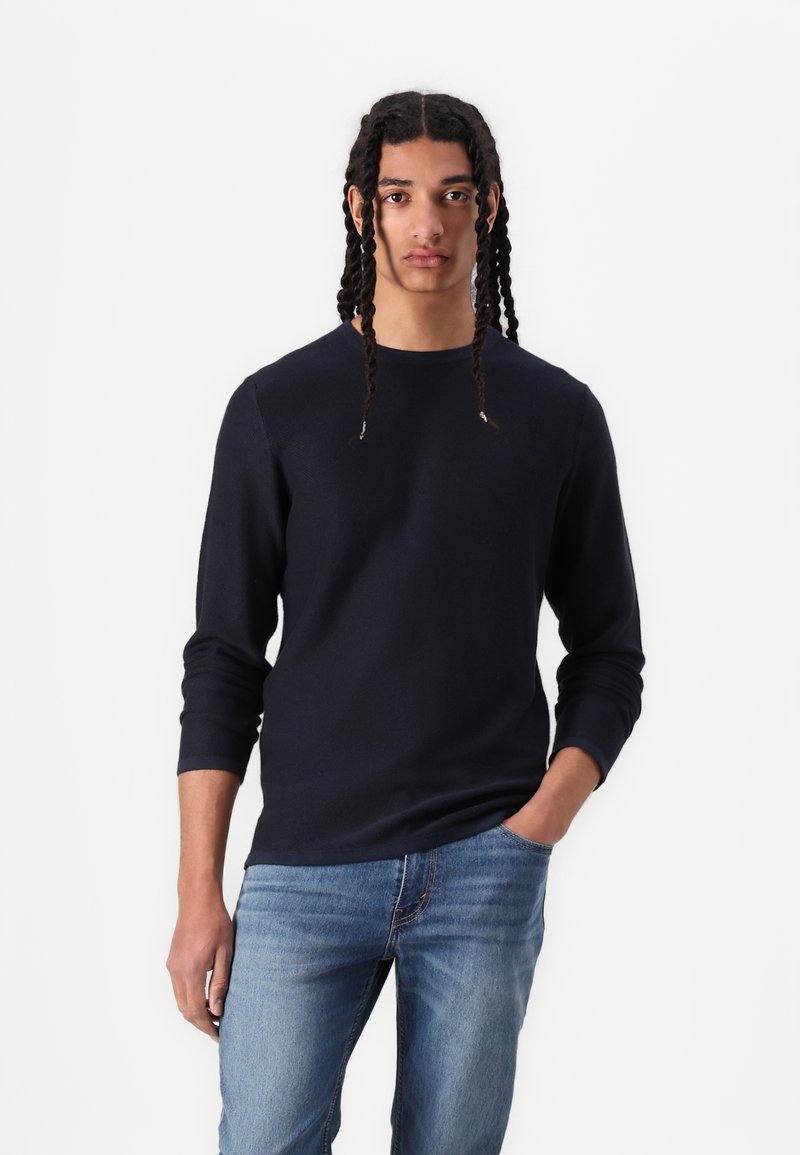 Marineblaue Langarmbluse mit strukturiertem Strickmuster, Rundhalsausschnitt und gerippten Bündchen, kombiniert mit hellblauen Jeans und Händen in den Taschen.