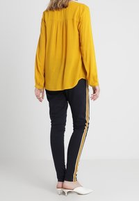 Haut à manches longues jaune moutarde avec des plis au dos, associé à un legging noir avec des rayures jaunes sur les côtés et des talons blancs.