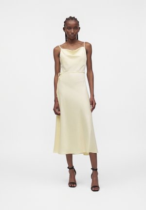 Vrouw draagt een bleekgele mouwloze midi-jurk met dunne bandjes en een zijstrikceintuur, gecombineerd met zwarte hakken met enkelriempje, op een effen achtergrond.