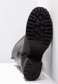 Botte noire avec semelle épaisse à crampons, fermeture éclair latérale visible, semelle tournée vers le haut sur fond blanc et mur, pointure marquée 37.