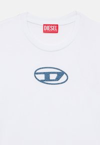 Λευκό t-shirt με στρογγυλή λαιμόκοψη, με κόκκινη ετικέτα μάρκας DIESEL και μπλε οβάλ λογότυπο με στυλιζαρισμένο «D» στο κέντρο στο στήθος.