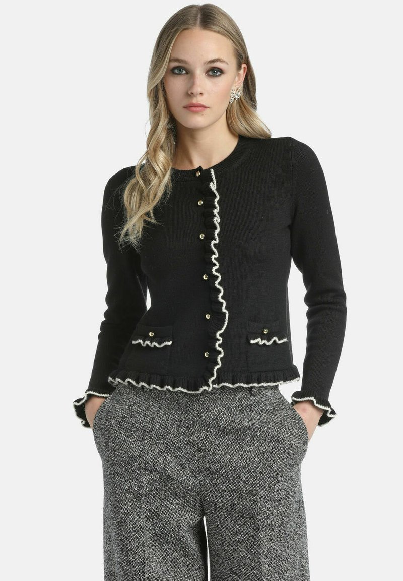 Melna džemperis ar baltām volānu akcentiem, pogu priekšpuse un divas kabatas. Izgatavots no teksturētas adījuma auduma, pieguļošs modelis.