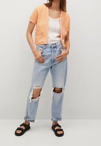 Pfirsichfarbener, gestrickter, gekürzter Cardigan mit kurzen Ärmeln, über einem weißen Tanktop getragen. Hellblaue, distressed gerade Jeans und schwarze Sandalen.