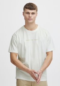 Lys beige bomull T-skjorte med korte ermer og rund hals. Har brodert tekst "HVA ER DET SOM HASTER?" i kontrastfarge gull.