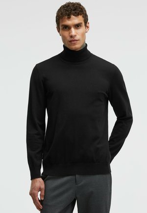 Black turtleneck sweater made of smooth knit fabric, featuring long sleeves and a ribbed collar and hem, worn with grey trousers.