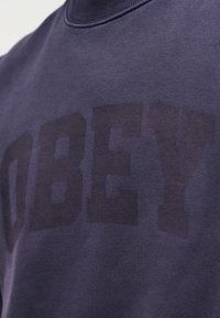 Σκούρο μπλε φούτερ με στρογγυλή λαιμόκοψη από απαλό ύφασμα, με μεγάλο, τονικό τύπωμα "OBEY" στο μπροστινό μέρος. Λεια υφή και απλός σχεδιασμός.