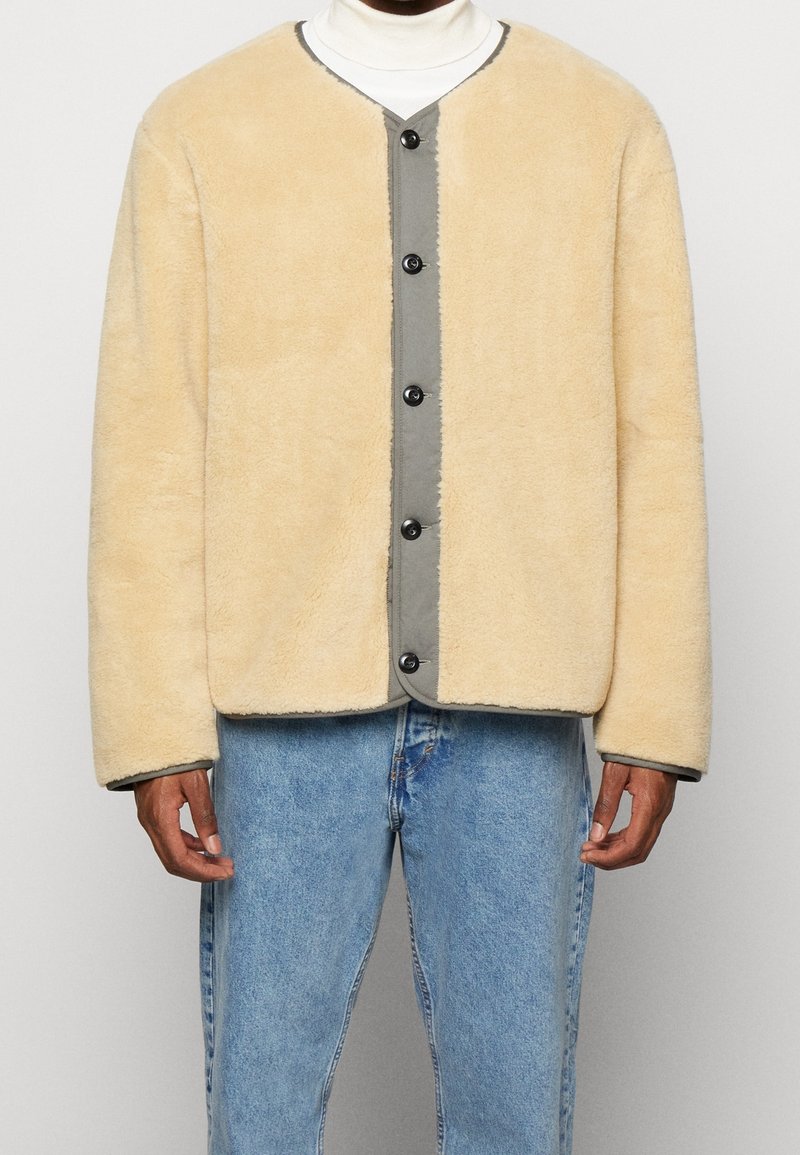 Giacca fuzzy beige con una chiusura frontale grigia a bottoni, dotata di un collo rotondo e bottoni neri. Indossata sopra un dolcevita bianco e jeans in denim.