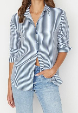 Frau trägt ein blau-weiß vertikal gestreiftes Hemd mit hochgekrempelten Ärmeln, kombiniert mit hellblauen Jeans, Hand in der Tasche.