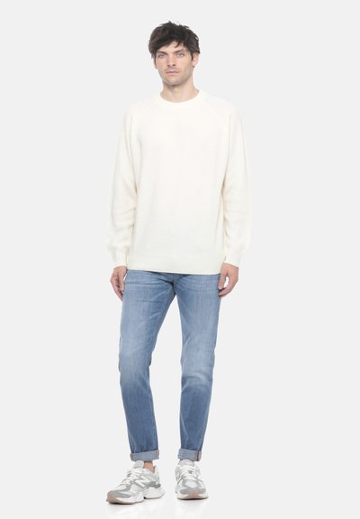 Uomo in piedi che indossa un maglione bianco a maniche lunghe, jeans azzurri con risvolti e sneaker bianco-grigie su uno sfondo bianco uniforme.