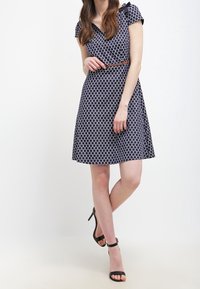 Vestido azul marino con lunares blancos, mangas cortas, escote en V y cintura entallada, acentuado por un cinturón marrón. Sandalias de tacón negro.