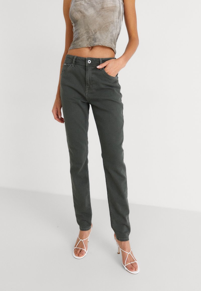 Pantalones de corte slim en verde oliva con una textura suave, que cuentan con un cierre frontal de botón y detalles de costura sutiles. Llevados con un top corto y tacones blancos.
