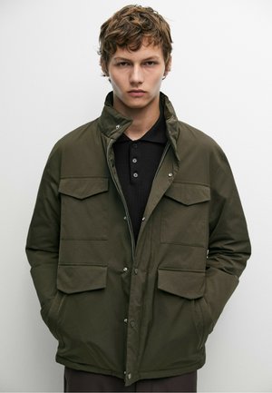 Ung man med kort lockigt hår som bär en olivgrön jacka över en svart knäppt skjorta, stående mot en enfärgad vit bakgrund.
