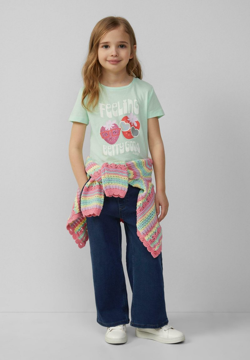 Muntgroene t-shirt met de tekst "Feeling Berry Good" en fruitillustraties; gecombineerd met wijde blauwe jeans en een meerkleurige gebreide sjaal.