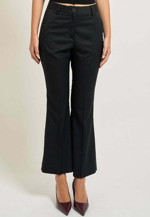 Donna che indossa pantaloni neri svasati corti e tacchi a spillo rossi scuri, in piedi davanti a uno sfondo semplice.