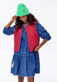 Červená prošívaná vesta s modrými akcenty přes džínové šaty s dlouhým rukávem, které mají vrstvenou sukni a vyšívané detaily, doplněné zelenou pletenou čepicí.