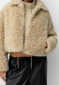 Beige faux fur cropped jakke med en blød tekstur, knaplukning og sidelommer, parret med sorte bukser med snøre.