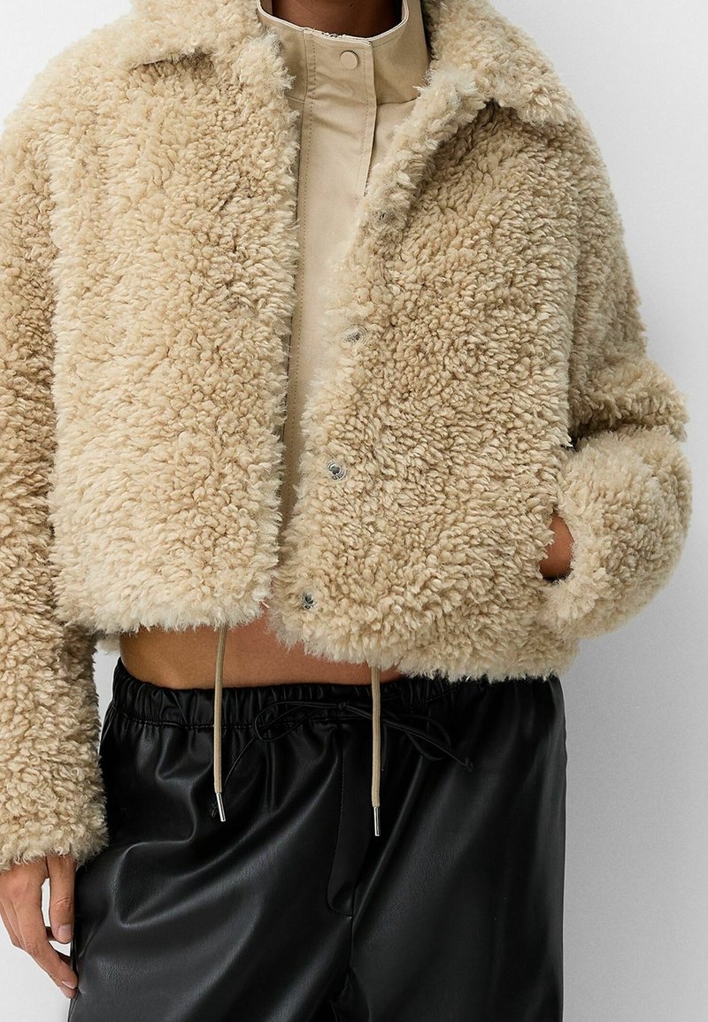 Beige faux fur cropped jakke med en blød tekstur, knaplukning og sidelommer, parret med sorte bukser med snøre.