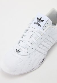 Fehér bőr sneaker három csíkkal, texturált részletekkel, párnázott nyelvvel, valamint fekete Adidas logóval a nyelven és az oldalpaneleken.