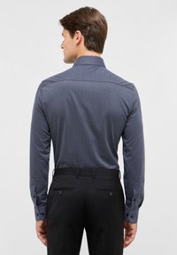Langarm overhemd met knopen in marineblauw met een geometrisch patroon; beschikt over een klassieke kraag en contrasterende knoopmanchetten. Gepaard met zwarte broeken.