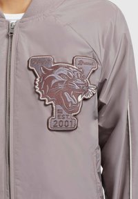Veste bomber rose clair avec un grand écusson brodé représentant un tigre en marron, accompagné du texte "Khyja Brand" et "EST 2001." Matière lisse.