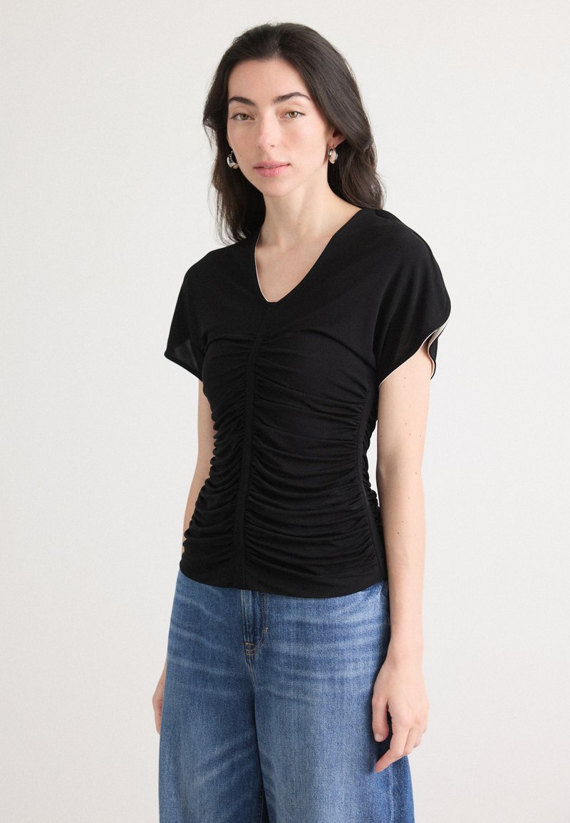 Black short-sleeve top made from soft fabric, featuring gathered texture along the front and a v-neckline, paired with light blue denim jeans.